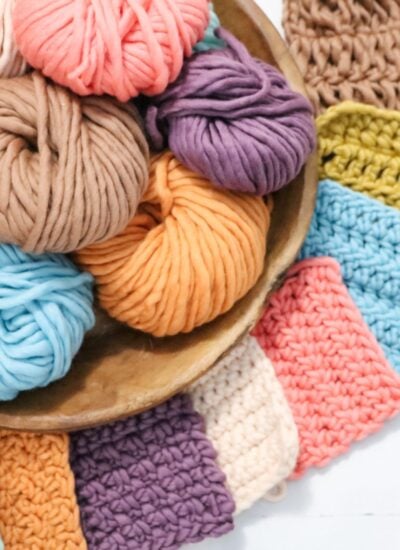 A wooden bowl filled with colorful skeins of yarn, surrounded by crocheted swatches in various pastel and bright colors.