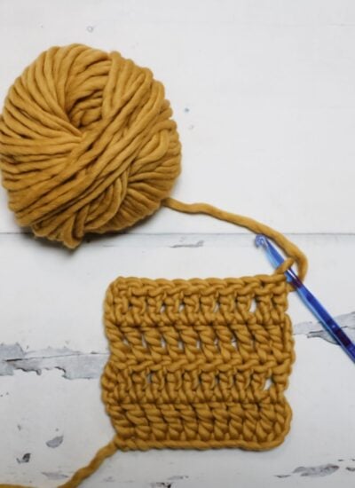 A ball of thick mustard yellow yarn and a blue crochet hook attached to a square swatch of crocheted fabric on a white surface.