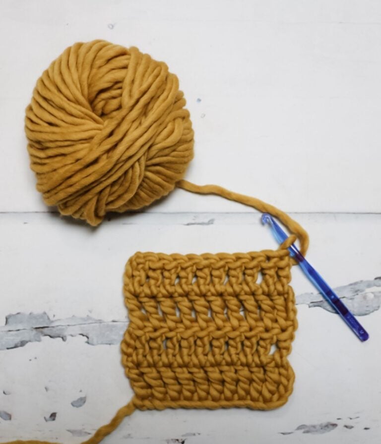 A ball of thick mustard yellow yarn and a blue crochet hook attached to a square swatch of crocheted fabric on a white surface.