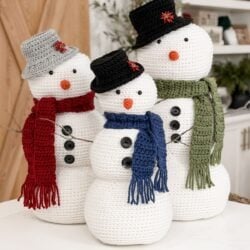 Three Crochet Snowman figures wear hats, scarves, and buttons, standing together indoors. Each snowman features a carrot nose and different colored accessories: red, blue, and green.