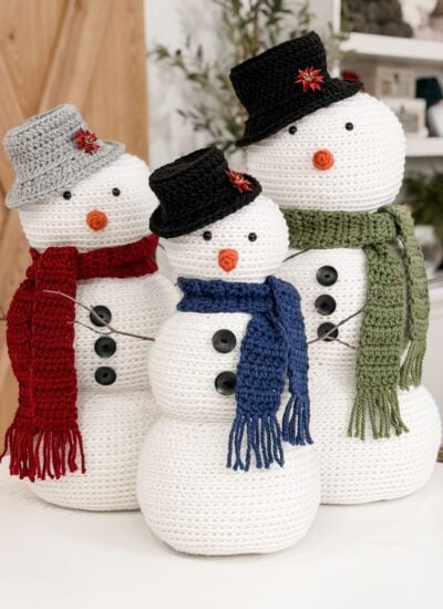 Three Crochet Snowman figures wear hats, scarves, and buttons, standing together indoors. Each snowman features a carrot nose and different colored accessories: red, blue, and green.