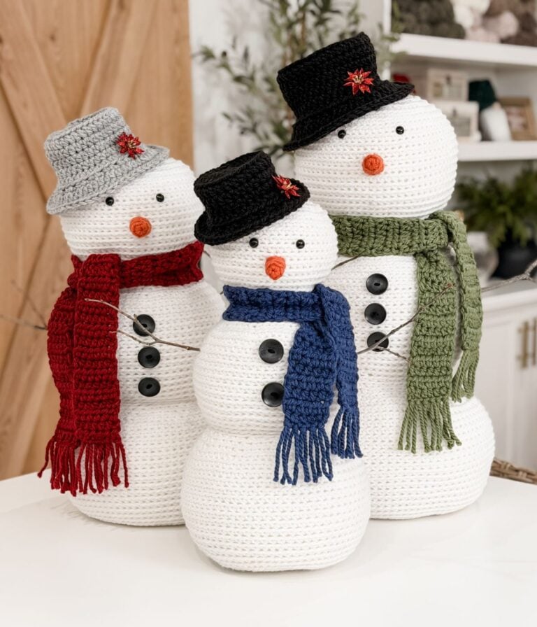 Three Crochet Snowman figures wear hats, scarves, and buttons, standing together indoors. Each snowman features a carrot nose and different colored accessories: red, blue, and green.