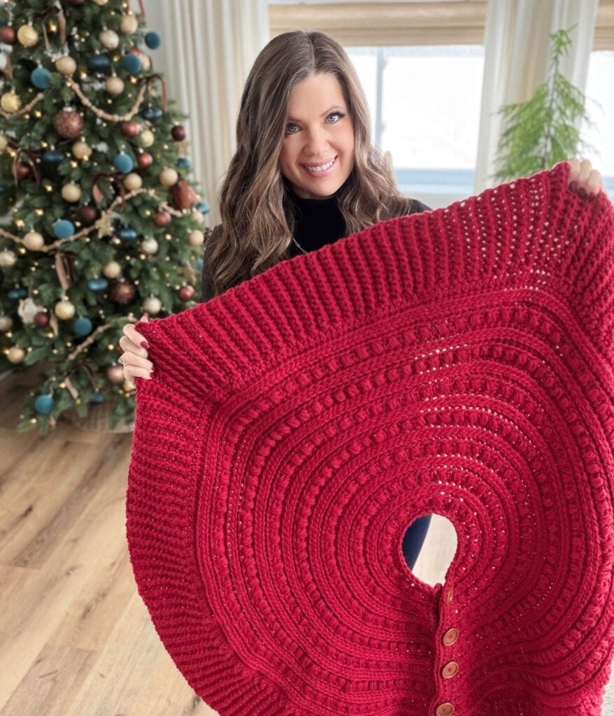 Woman holding a large red crocheted tree skirt in a living room with a decorated Christmas tree and a charming crochet snowman in the background.
