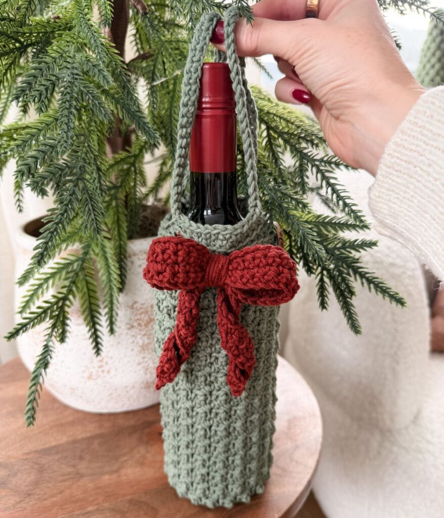 A hand holds a bottle of wine in a green crocheted wine cozy with a red bow, placed on a wooden surface near a potted plant.