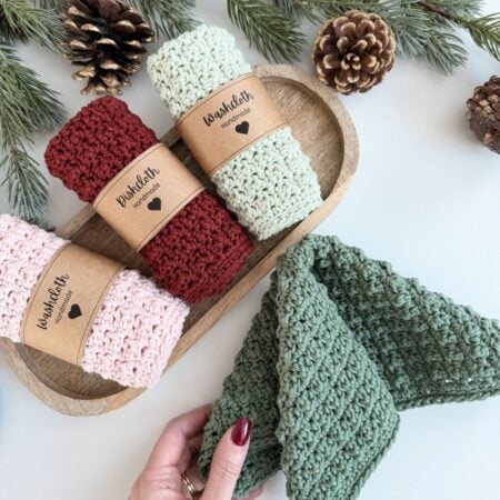 Three rolled knitted dishcloths in red, cream, and pink sit on a wooden tray, with a hand holding a green dishcloth. Pinecones and evergreen branches are in the background.