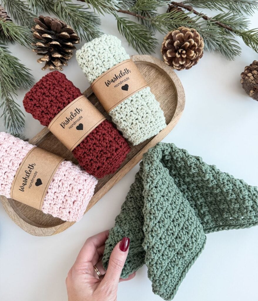 Three rolled knitted dishcloths in red, cream, and pink sit on a wooden tray, with a hand holding a green dishcloth. Pinecones and evergreen branches are in the background.