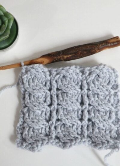 A wooden crochet hook and a swatch of light gray textured crochet work are placed on a white surface next to a small potted succulent.