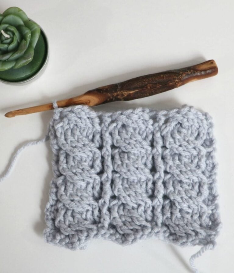 A wooden crochet hook and a swatch of light gray textured crochet work are placed on a white surface next to a small potted succulent.