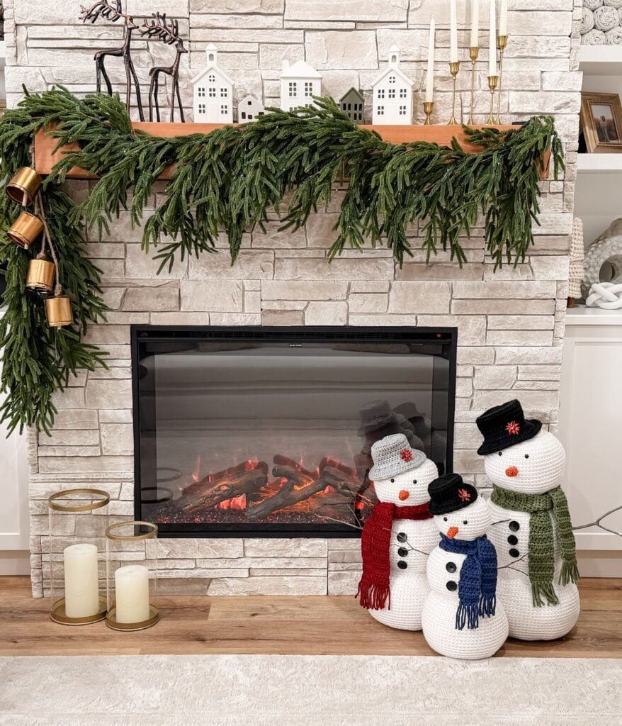 A decorated fireplace features a garland, gold bells, and miniature white houses on the mantle. Three crochet snowmen with scarves and hats stand cheerfully in front of the fireplace.