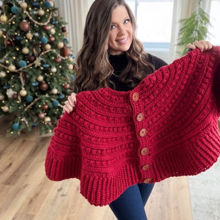 A woman smiles while holding up a large, red, hand-knitted sweater with wooden buttons. A crochet snowman ornament hangs on the decorated Christmas tree in the background.