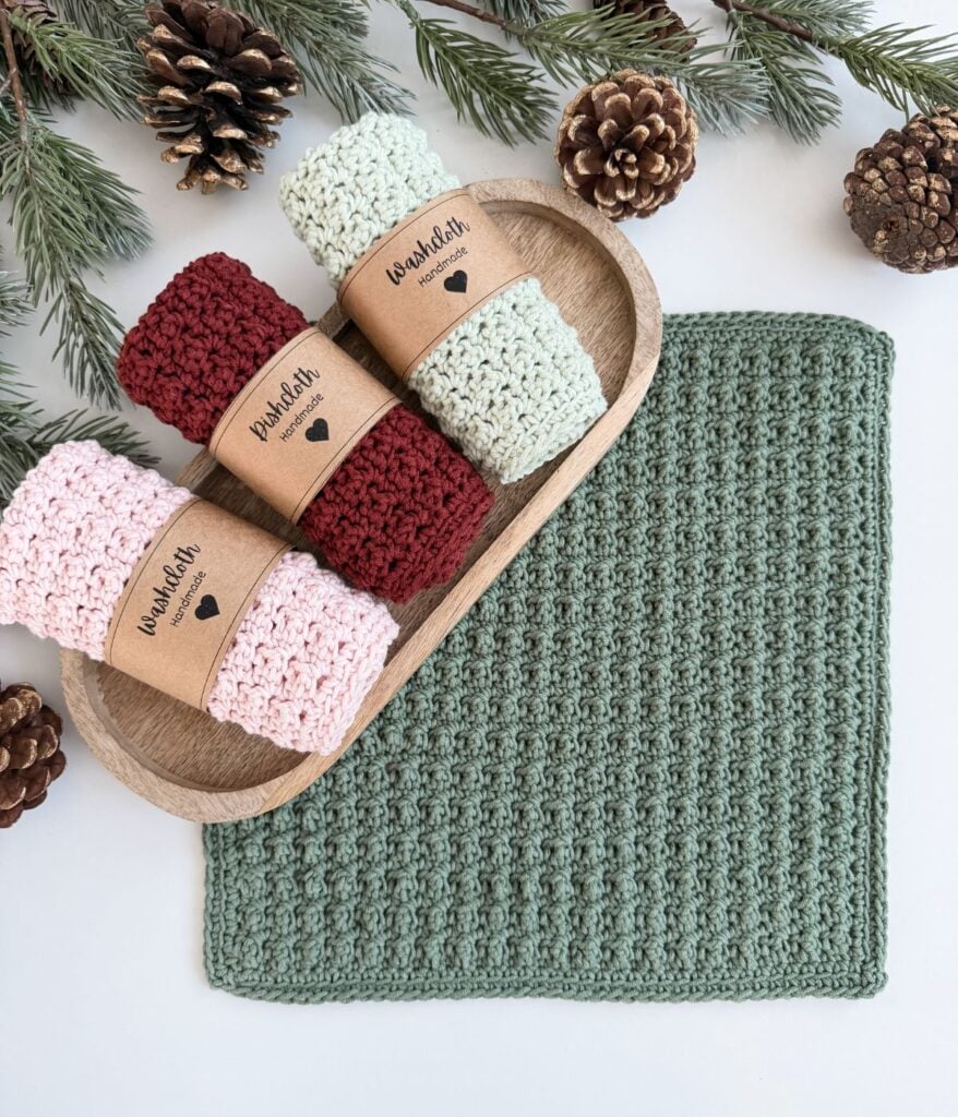 A green crocheted dishcloth lies next to a wooden tray holding four rolled washcloths in red, pink, and cream, with pinecones and evergreen branches nearby.