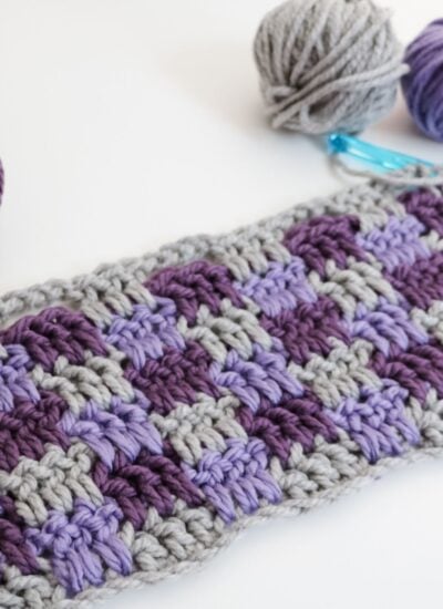 Crocheted swatch in a checkerboard pattern using gray, purple, and lavender yarn, with two yarn balls and a blue crochet hook nearby on a white surface.