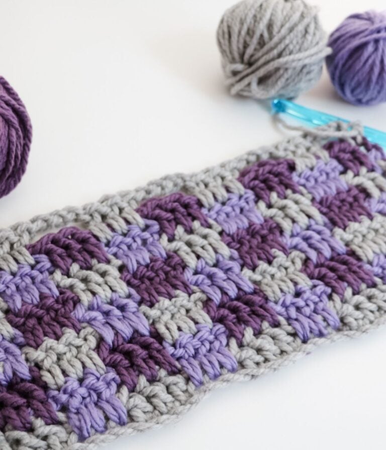 Crocheted swatch in a checkerboard pattern using gray, purple, and lavender yarn, with two yarn balls and a blue crochet hook nearby on a white surface.