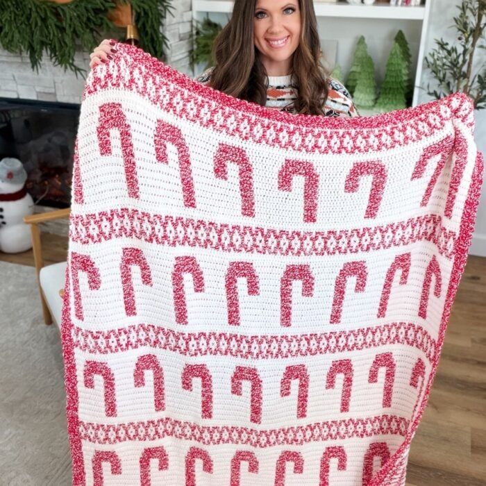 A woman holds up a large mosaic blanket featuring red and white candy cane patterns in a living room decorated for the holidays.