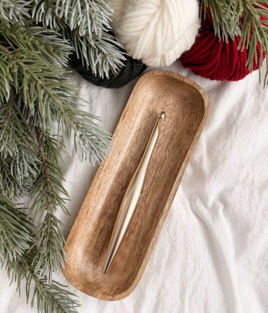 A metal crochet hook rests in a wooden tray, surrounded by pine branches and yarn balls in white, red, and green—perfect for crafting a festive Crochet Snowman—on a white fabric background.