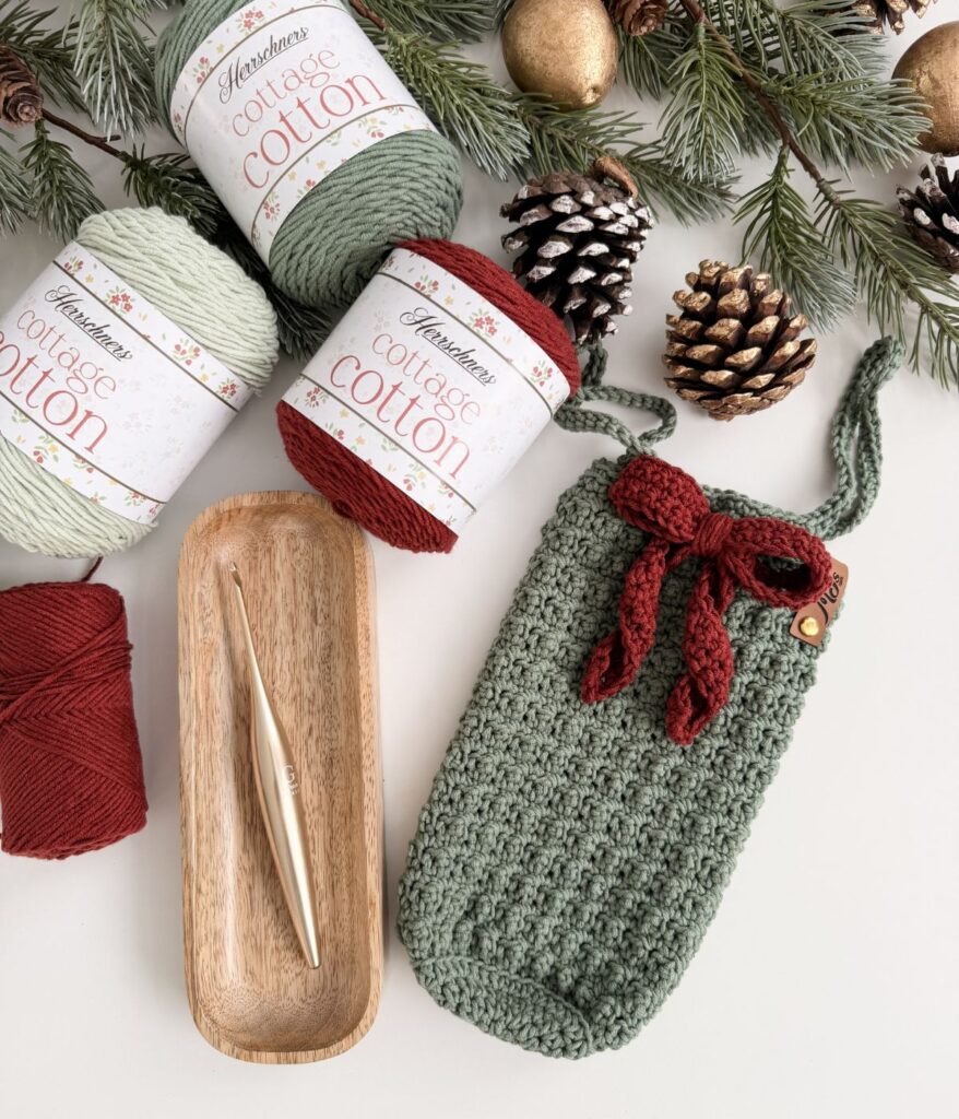 Three skeins of cottage cotton yarn, a wooden tray with a crochet hook, and a green crocheted wine cozy tied with a red bow are arranged on a white surface with pinecones and holiday decor.