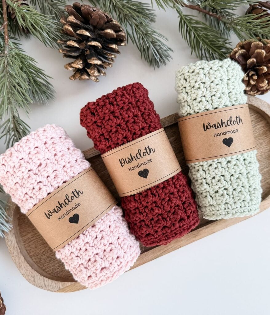 Three rolled, hand-crocheted washcloths and dishcloths in pink, red, and light green with handmade labels, placed on a wooden tray, surrounded by pine branches and pinecones.