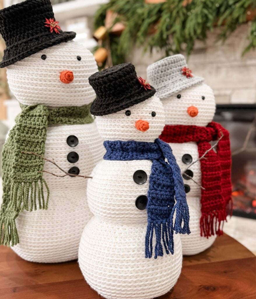 Three Crochet Snowman figures with black hats and scarves in green, blue, and red stand on a wooden surface in front of a fireplace and greenery.