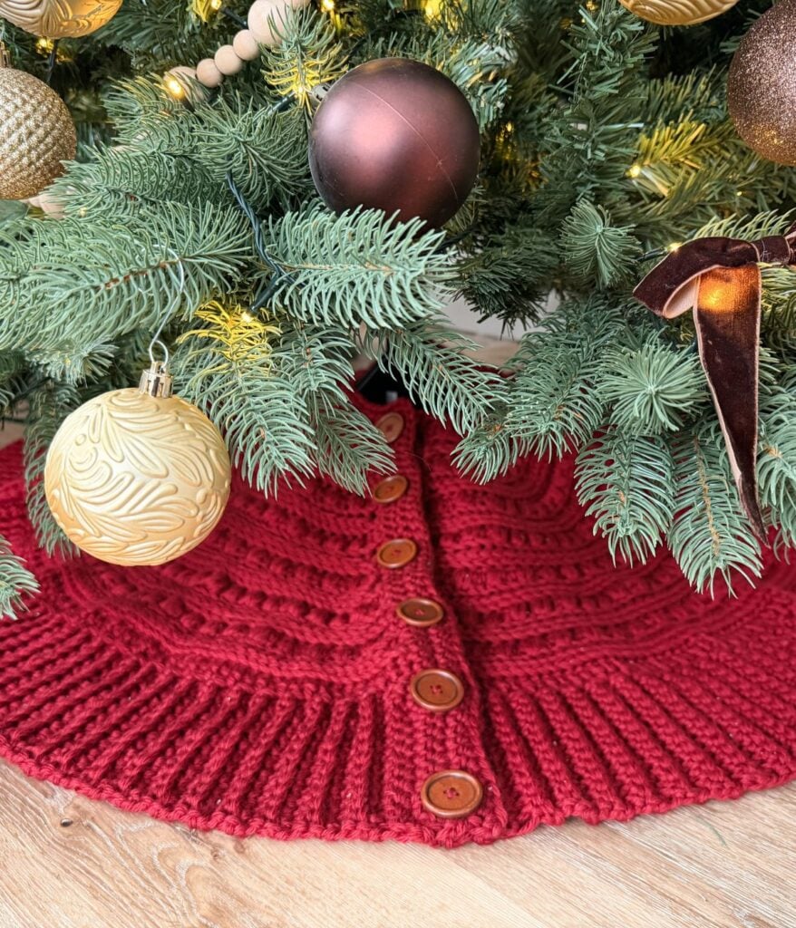A red knitted tree skirt with wooden buttons is arranged under a decorated Christmas tree with gold and brown ornaments, complemented by a charming crochet snowman nearby.