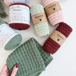 Various handmade crocheted washcloths and dishcloths in green, pink, and red are displayed with labeled yarn spools and pinecones on a white surface. A hand holds a green washcloth in the corner.