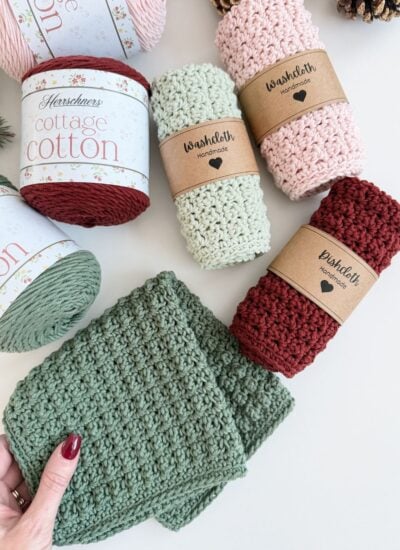 Various handmade crocheted washcloths and dishcloths in green, pink, and red are displayed with labeled yarn spools and pinecones on a white surface. A hand holds a green washcloth in the corner.