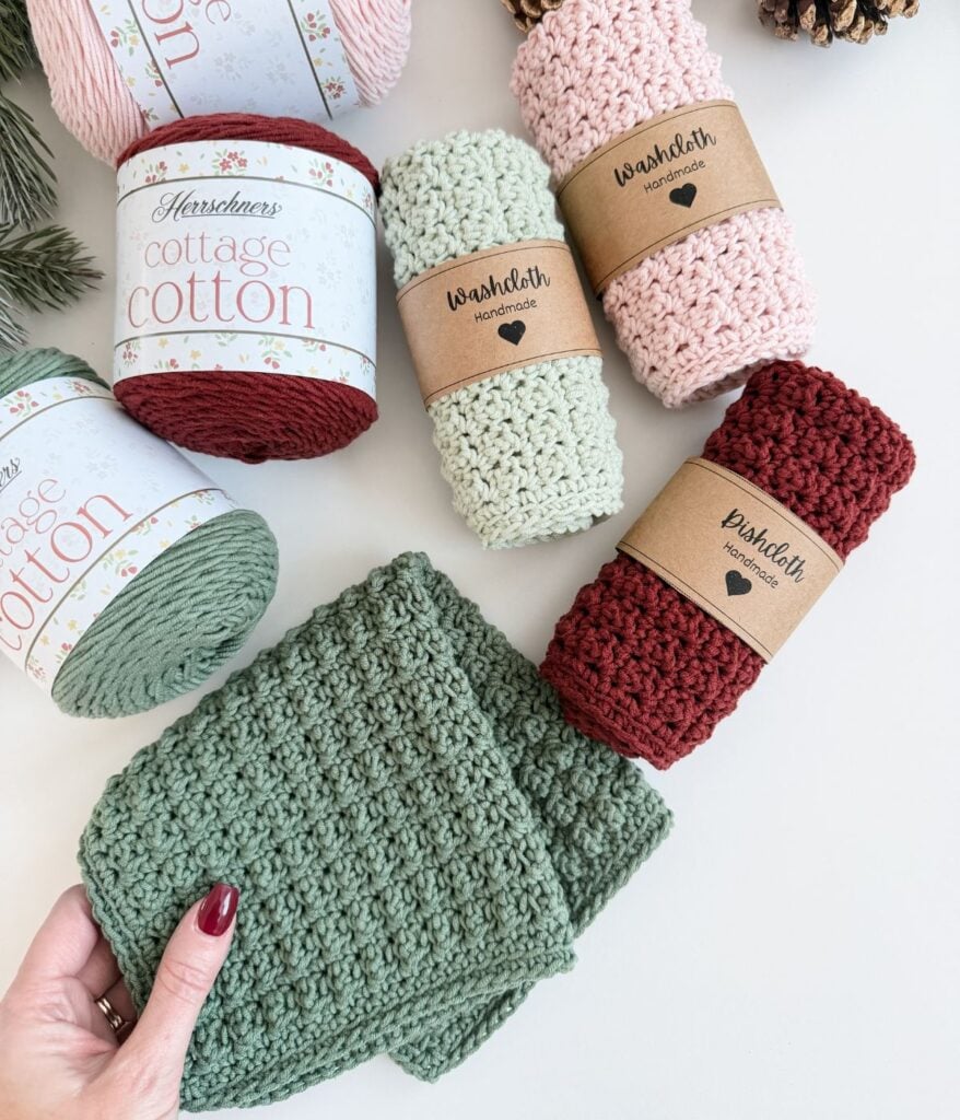 Various handmade crocheted washcloths and dishcloths in green, pink, and red are displayed with labeled yarn spools and pinecones on a white surface. A hand holds a green washcloth in the corner.