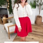 Woman wearing a white crocheted cardigan and red skirt stands indoors, smiling, with holiday decorations and greenery in the background. Text reads "WINTER SPARK CARDIGAN Crochet Pattern & Video—create your perfect winter cardigan!.