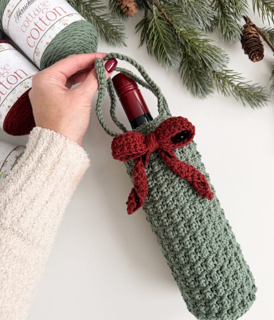 A hand holds a green crocheted Wine Cozy with a red bow, containing a wine bottle; yarn skeins and pine branches are in the background.