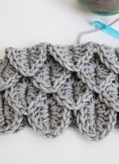 A section of gray crocheted fabric featuring a textured crocodile stitch pattern, with a blue crochet hook and yarn visible in the background.