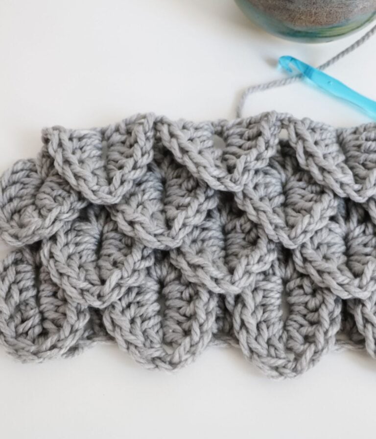 A section of gray crocheted fabric featuring a textured crocodile stitch pattern, with a blue crochet hook and yarn visible in the background.