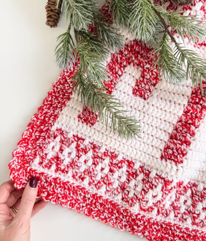 A hand holds the corner of a red and white mosaic blanket with a festive crocheted pattern, partially covered by pine branches.