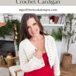 A woman wearing a white crocheted cardigan over a red dress stands indoors, smiling at the camera. Shelves with decor and a fireplace are in the background. Text promotes this lovely cardigan crochet pattern.