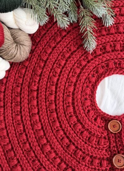 Close-up of a red, crocheted tree skirt with wooden buttons, surrounded by yarn skeins, pine branches, and a cute crochet snowman for a festive touch.