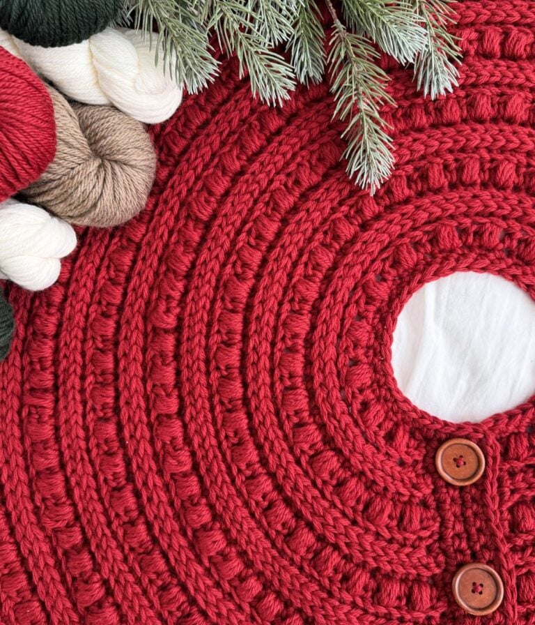 Close-up of a red, crocheted tree skirt with wooden buttons, surrounded by yarn skeins, pine branches, and a cute crochet snowman for a festive touch.