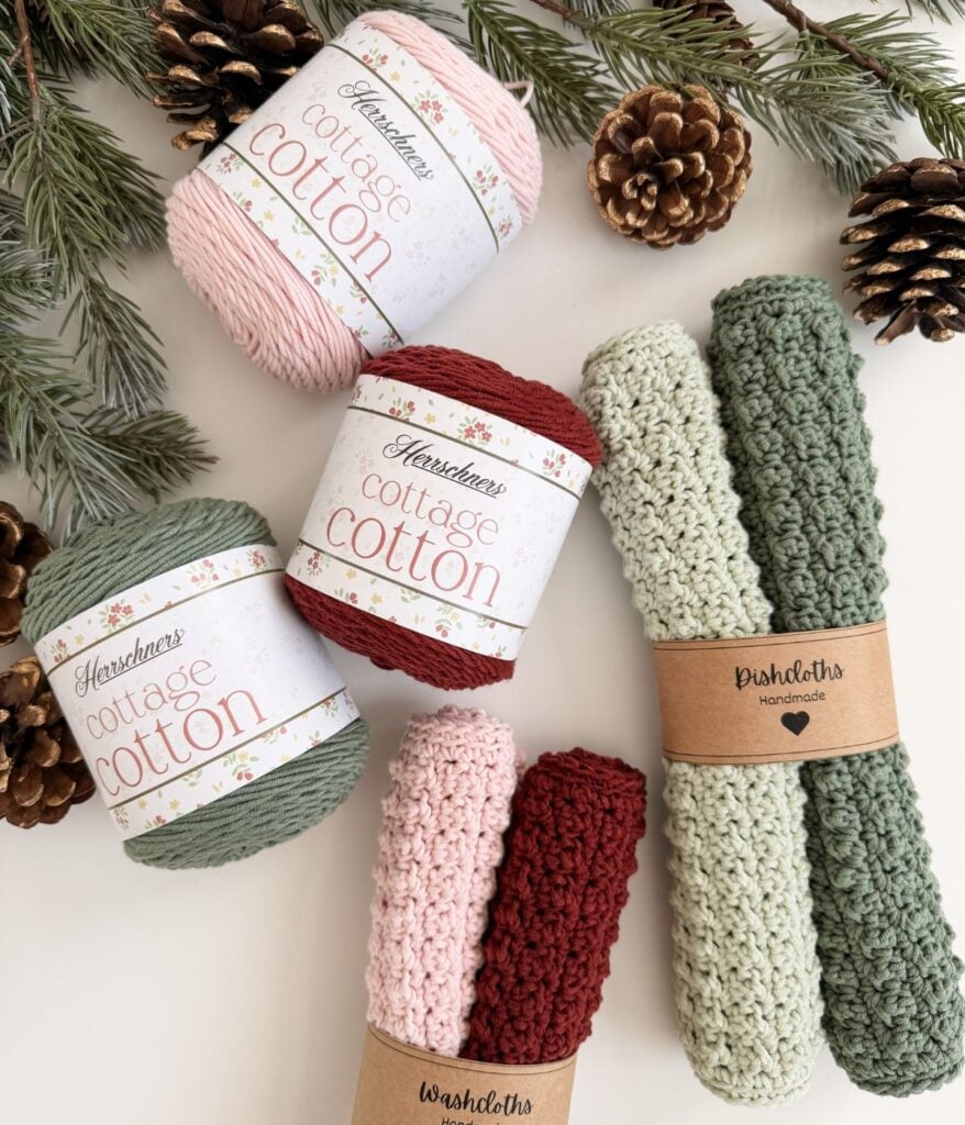 Three skeins of cottage cotton yarn in pink, red, and green are arranged with pinecones, fir branches, and two rolled, handmade dishcloths—perfect for adding a cozy touch to your kitchen or gifting a cheerful dishcloth set.