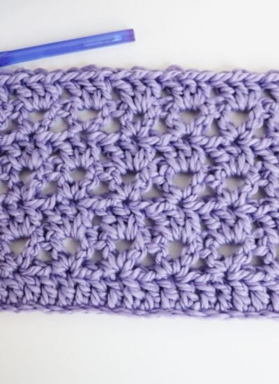 A rectangular swatch of lilac-colored crocheted fabric with an open, lacy pattern is shown beside a purple crochet hook on a white background.