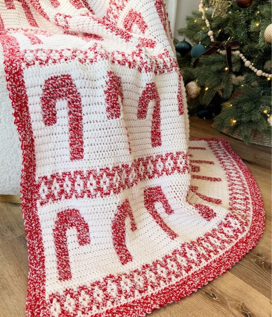 A mosaic blanket with red and white candy cane patterns is draped near a decorated Christmas tree on a wooden floor.