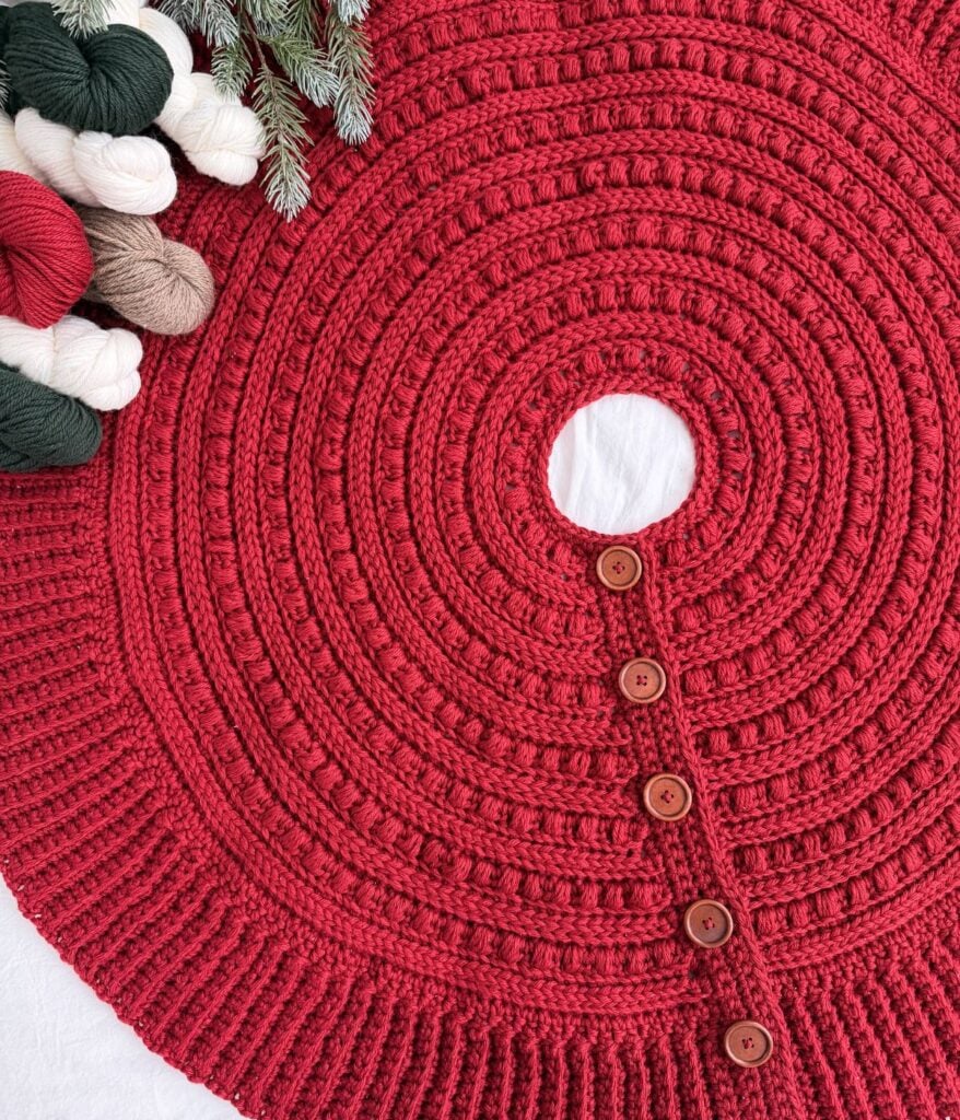 A round, red knit tree skirt with brown buttons is displayed flat, surrounded by pine branches, skeins of yarn in festive hues, and a charming crochet snowman for a cozy holiday touch.