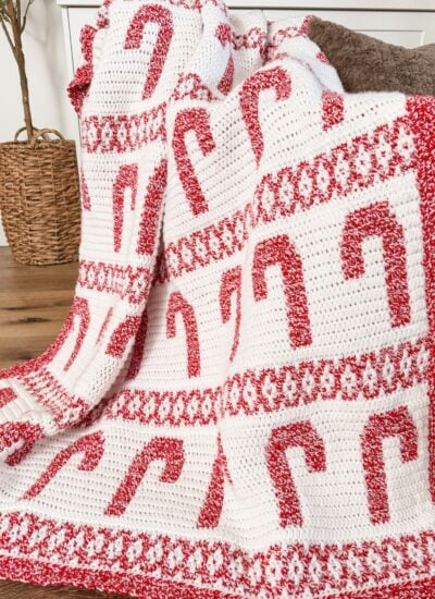 A crocheted mosaic blanket featuring red and white candy cane patterns is draped over a wooden rod in a cozy living room setting.