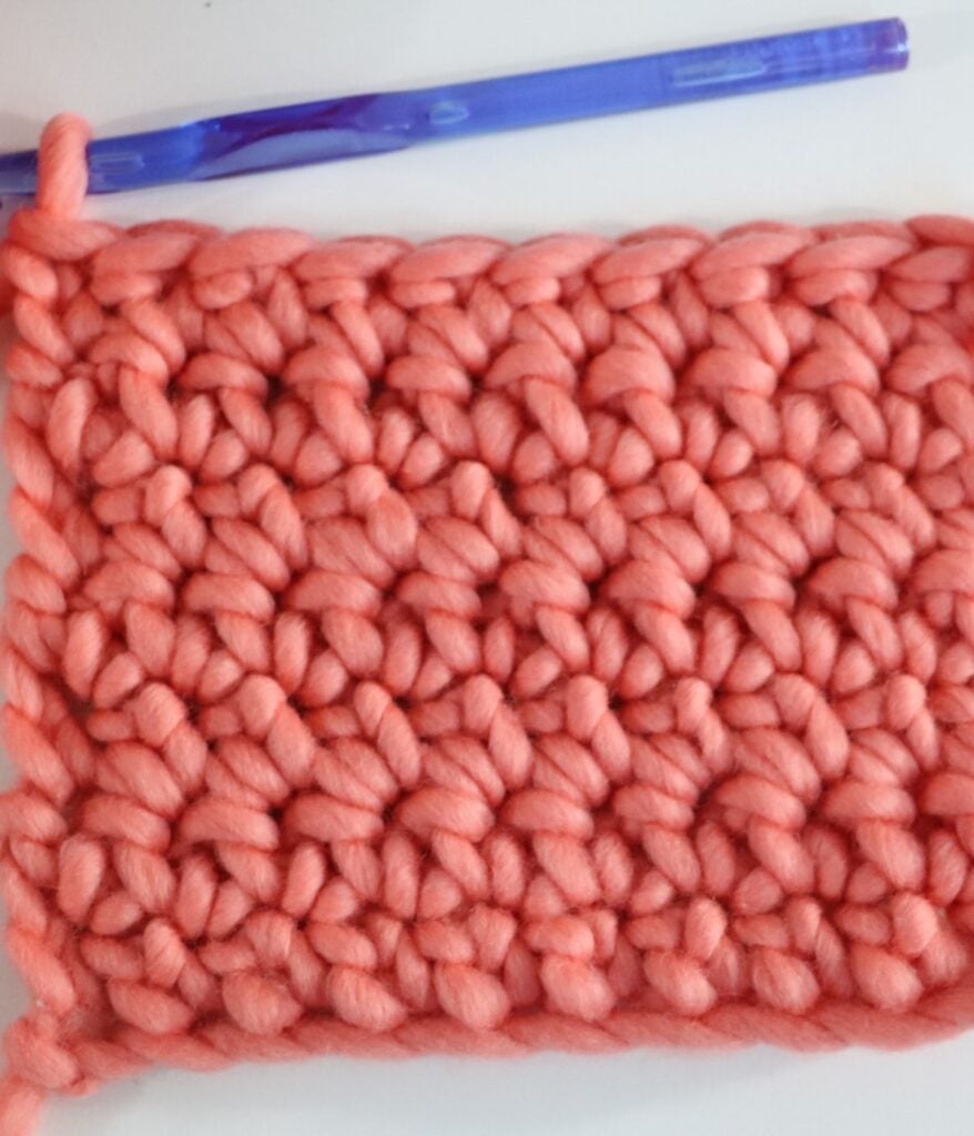 A swatch of chunky pink crochet knitting with a large blue crochet hook resting above it on a white surface.