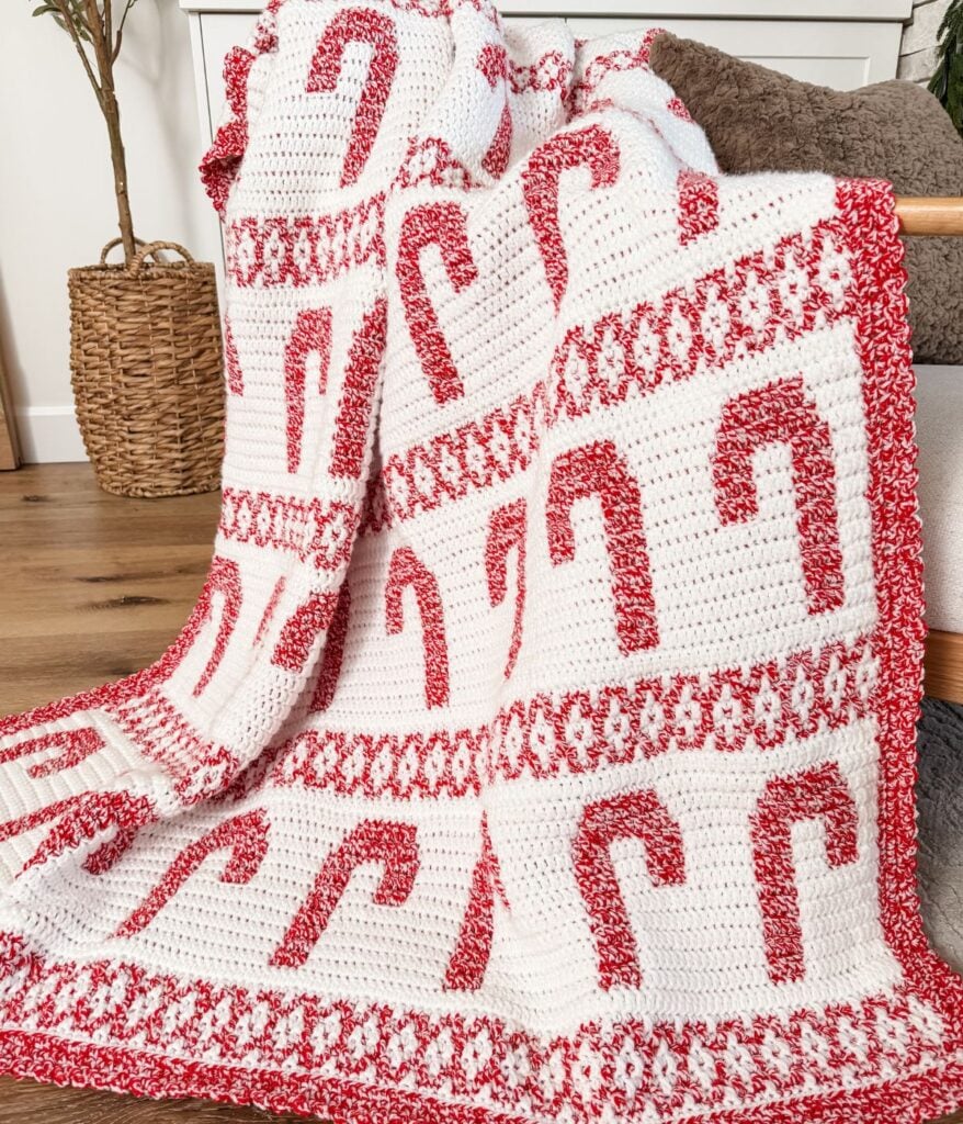 A crocheted mosaic blanket featuring red and white candy cane patterns is draped over a wooden rod in a cozy living room setting.