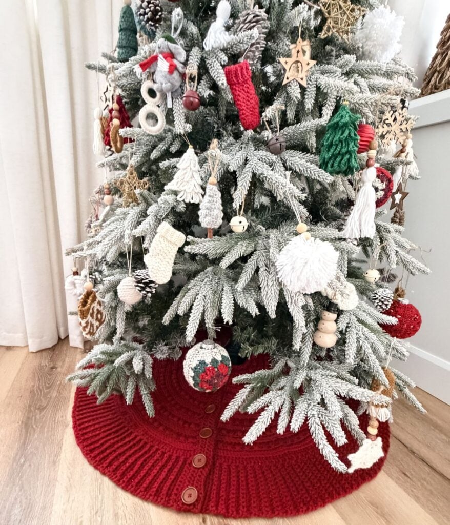 A Christmas tree with white-tipped branches is decorated with yarn ornaments and a crochet snowman, and sits on a red knitted tree skirt with buttons.