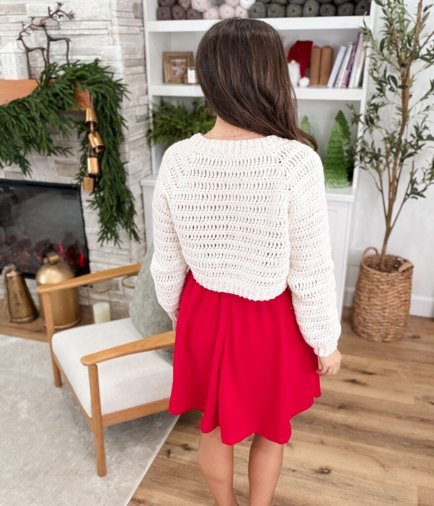 A woman with long brown hair, wearing a white knit cardigan and red skirt, stands with her back to the camera in a cozy, decorated living room.