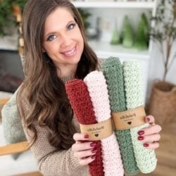 A woman smiles while holding four rolled, knitted dishcloths in pink, green, and red tones, each dishcloth neatly wrapped with a brown paper label.