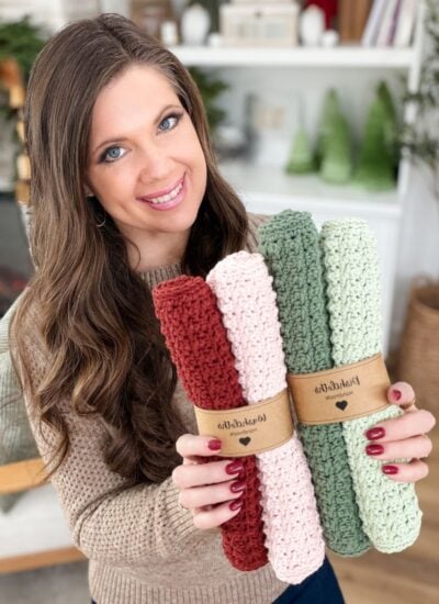 A woman smiles while holding four rolled, knitted dishcloths in pink, green, and red tones, each dishcloth neatly wrapped with a brown paper label.