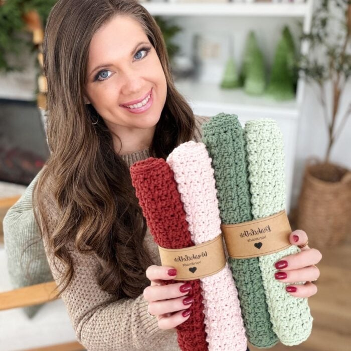 A woman smiles while holding four rolled, knitted dishcloths in pink, green, and red tones, each dishcloth neatly wrapped with a brown paper label.