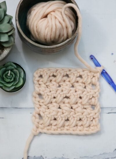 A crochet square made with chunky beige yarn, a blue crochet hook, a ball of yarn in a bowl, and two succulent plants on a white surface.