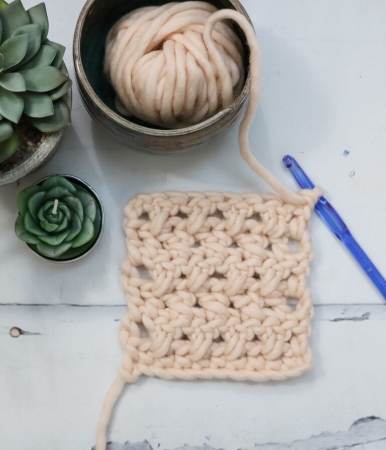 A crochet square made with chunky beige yarn, a blue crochet hook, a ball of yarn in a bowl, and two succulent plants on a white surface.