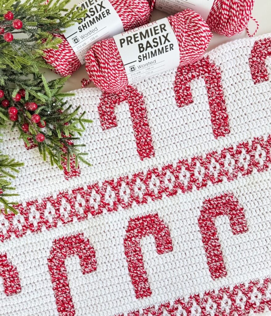 A crocheted white mosaic blanket with red candy cane and decorative patterns, displayed beside sprigs of greenery, red berries, and skeins of Premier Basix Shimmer yarn.