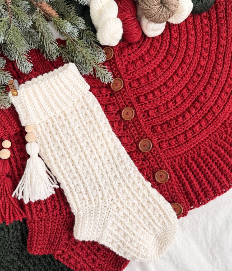A white knitted Christmas stocking with a tassel lies on a red textured blanket with brown buttons, surrounded by yarn skeins, pine branches, and a cozy tree skirt.